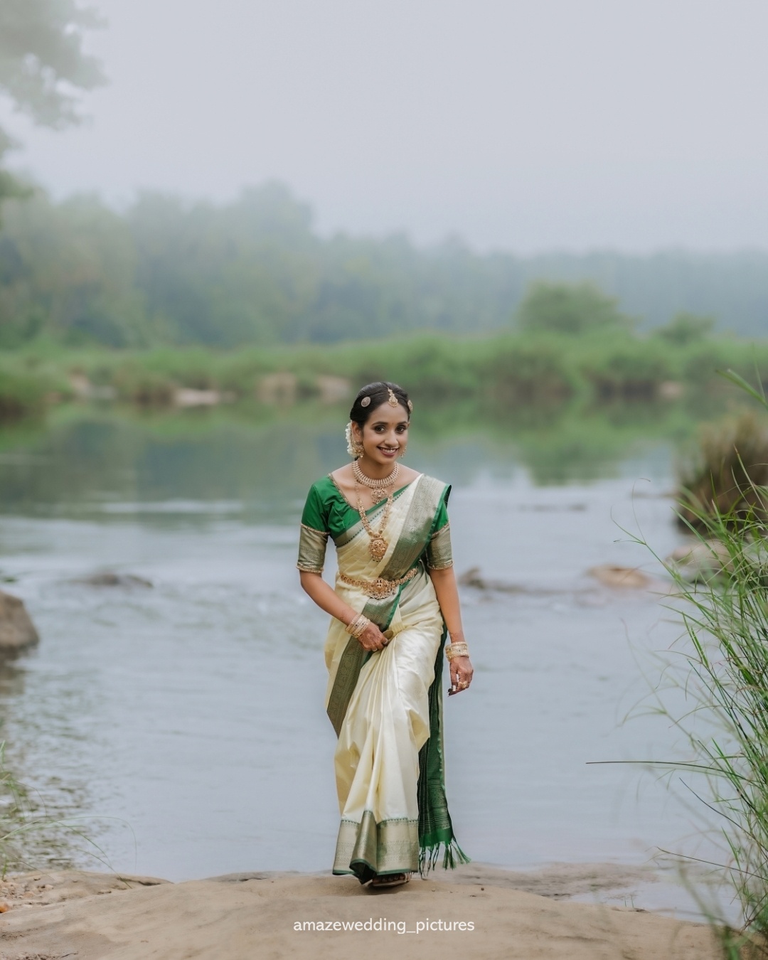 A bride in an ivory sari standing beside a riverside landscape